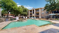The Aidan is a residential community whose standout amenity is its expansive, resort-inspired outdoor pool area. The centerpiece is a generously sized swimming pool with clear turquoise water, complemented by an adjacent circular spa, creating a relaxing retreat for residents. The pool deck is finished in warm, natural-toned flagstone pavers that extend across a broad sunbathing area furnished with contemporary chaise lounges and coordinating blue accent cushions, alongside multiple patio umbrellas providing ample shade throughout the day. A pergola structure at the rear of the pool area adds architectural character and additional shaded gathering space. The community is framed by mature deciduous trees that provide a lush, park-like ambiance and natural privacy. A wrought iron perimeter fence encloses the pool area for security and safety. The leasing office entrance is accessible via a covered stone-accented entryway with double glass doors, decorative lantern lighting, and manicured landscaping. The surrounding multi-story residential buildings feature a mix of stucco and natural limestone exteriors with private balconies, reflecting a classic Texas architectural style. The entire outdoor environment benefits from abundant natural sunlight, making it an inviting communal space designed for leisure and relaxation.