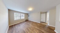 This single-floor unit offers a practical and clean layout designed for comfortable everyday living. The main living area is bright and open, featuring warm wood-look plank flooring, neutral beige walls, and a large window that allows natural light to fill the space. A wall-mounted HVAC unit provides climate control, and a pass-through window connects the living area directly to the kitchen, enhancing the open feel of the space.

The kitchen is thoughtfully designed with white shaker-style cabinetry extending to the ceiling for generous storage, light granite-style countertops, a stainless steel sink, and a full suite of black appliances including a refrigerator, dishwasher, and range with overhead ventilation. The wood-look flooring continues seamlessly into this space.

The bedroom offers plush carpeting for comfort underfoot, a ceiling fan with light fixture, a window with blinds, and ample wall space. The room is calm and well-proportioned.

The bathroom is clean and functional, featuring a white vanity with a granite-style countertop, a large mirror with bright vanity lighting, and a full soaking tub and shower combo with a white surround. Wood-look flooring ties the bathroom's aesthetic to the rest of the unit. Overall, the space presents a cohesive, updated aesthetic throughout.