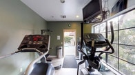 This compact yet fully equipped fitness center offers everything needed for a well-rounded workout routine. The room is finished with sage green walls and light-toned flooring, creating a clean, focused atmosphere enhanced by recessed lighting throughout. A large wall-spanning mirror runs along one side of the space, visually expanding the room and allowing users to monitor their form during exercise. Natural light filters in through a sizable window framing views of the surrounding greenery, adding an inviting, open feel to the interior. The cardio section is well-appointed, featuring multiple treadmills from Star Trac, a recumbent bike, an upright stationary bike, and an elliptical trainer. A wall-mounted flat-screen television is positioned for easy viewing from the cardio equipment. The strength training area includes a multi-function weight machine with adjustable seating, a pull-up and dip station, and a traditional standing scale. Rubber-matted flooring sections are placed beneath key equipment for safety and durability. A direct door provides seamless access to an outdoor area where a pool and patio seating are visible, offering a convenient transition between indoor and outdoor fitness and leisure activities. A wall clock and informational signage complete the functional, user-friendly design of the space.