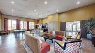 Located at 14689 Briar Forest Dr in Houston, TX, this single-floor community clubhouse offers a generously sized, open-plan layout designed for both socializing and productivity. The main gathering area is anchored by warm golden-yellow walls, high ceilings with recessed lighting, and large windows dressed in bold red and orange drapes that flood the space with natural light and views of the surrounding grounds. Comfortable seating arrangements — including leather sofas, patterned accent chairs, and a central coffee table — provide inviting areas for relaxation, while a wall-mounted large-screen TV adds an entertainment focal point. A full kitchen features dark cabinetry, a striking red brick mosaic tile backsplash, granite countertops, stainless steel appliances, and a built-in island with bar seating. A billiards table and shuffleboard table serve as standout recreational amenities, reinforcing the social character of the space. A dedicated business center offers individual workstations with dark wood desks, ergonomic seating, and pendant lighting. A quieter lounge alcove with contemporary artwork and slate tile flooring provides a more intimate retreat. Sliding barn doors and grey panel doors connect the various zones seamlessly throughout.