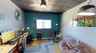 This single-floor space offers a well-organized and visually dynamic interior designed for both focused work and casual interaction. The most striking feature is a deep teal accent wall that anchors the room, creating a bold contrast against the lighter surrounding walls and dark drop-ceiling tiles. A ceiling fan provides air circulation overhead, contributing to a comfortable working environment.

The layout is thoughtfully divided into distinct functional zones. A dedicated workstation along one wall features a desk with dual monitors, a task lamp, and an ergonomic office chair — ideal for extended productivity sessions. Adjacent to this is a compact storage cabinet, keeping the workspace tidy and organized.

At the center of the room, a tall bistro-style table paired with a red bar stool and floor lamp creates an informal collaboration or break area. A plush, oversized bean bag chair adds a relaxed lounge element, offering a comfortable alternative seating option. The space is further warmed by layered area rugs with teal geometric accents that tie together the room's color palette.

Natural light enters through a single window on the teal wall, brightening the interior. A large mirror on an adjacent wall enhances the sense of openness. Overall, this space combines functionality with a creative, energetic aesthetic.