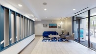 This single-floor lobby space presents a clean, contemporary design centered around comfort and functionality. Upon entry, automatic glass sliding doors open into a spacious, light-filled foyer with large-format tile flooring throughout. The entrance area immediately transitions into two distinct seating zones, each thoughtfully appointed to encourage relaxation and conversation.

The primary lounge area features a bold royal blue sofa paired with gray upholstered accent chairs, arranged around a light wood coffee table adorned with small potted plants. A vibrant patterned area rug in blue and white anchors the arrangement, while a trio of framed cityscape photographs adds an urban, cosmopolitan character to the neutral gray walls. Pendant lighting with cylindrical shades hangs above, complementing the recessed ceiling lights to create a warm, layered ambiance.

A secondary seating nook near the entrance includes two tan armchairs with mustard yellow cushions and a compact side table, positioned alongside a well-stocked open bookshelf — a thoughtful touch for visitors. A decorative blue panel wall system adds a striking architectural accent along one side of the lobby, introducing a pop of color that ties the space together. Floor-to-ceiling windows along the storefront invite natural light and provide views of the surrounding streetscape, enhancing the open, airy feel of this welcoming communal space.