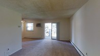 This single-floor space is built within a classic brick exterior structure and offers a practical, straightforward layout. The main living area is generously sized, featuring wall-to-wall carpeting underfoot and a textured ceiling overhead. A ceiling fan provides additional air circulation, while a wall-mounted air conditioning unit and baseboard heaters along the perimeter ensure year-round climate control. Natural light enters through a standard window on one wall, and a set of sliding glass doors fitted with vertical blinds opens directly onto a covered balcony, creating a seamless connection between the interior and exterior.

The covered balcony is a notable feature of this space, running the length of the unit's exterior and enclosed by decorative wrought iron railings. Constructed alongside the building's brick facade, the balcony offers a sheltered outdoor area with open views of a well-maintained green landscape, including mature evergreen trees and manicured shrubs. The concrete flooring and covered overhead structure make it a functional outdoor space suitable for a variety of uses regardless of weather conditions.