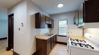 This single-floor unit offers a practical and straightforward layout, with warm hardwood flooring that runs consistently throughout the living areas and bedroom, lending a cohesive feel to the space. The kitchen is efficiently designed with dark espresso-toned cabinetry providing ample upper and lower storage, a light-toned countertop with a double-basin stainless steel sink, and a white gas range with a ventilation hood. A white refrigerator completes the appliance lineup, while a window above the sink area draws in natural light and offers views of surrounding greenery. A baseboard heating unit is also present in the kitchen area. The living area is open and airy, featuring a large window that fills the room with natural light, a ceiling fan, and a classic cast-iron radiator, adding a touch of vintage character. The bedroom is a quiet, well-lit room with a window framing outdoor tree views, light-toned hardwood floors, and dark wood baseboards. The bathroom is finished with white square wall tiles surrounding a full soaking tub and shower combination, complemented by a window for ventilation and a wall-mounted medicine cabinet. Overall, the space presents a clean, functional environment with classic details throughout.
