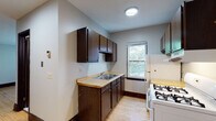 This single-floor unit offers a practical and straightforward layout, with warm hardwood flooring that runs consistently throughout the living areas and bedroom, lending a cohesive feel to the space. The kitchen is efficiently designed with dark espresso-toned cabinetry providing ample upper and lower storage, a light-toned countertop with a double-basin stainless steel sink, and a white gas range with a ventilation hood. A white refrigerator completes the appliance lineup, while a window above the sink area draws in natural light and offers views of surrounding greenery. A baseboard heating unit is also present in the kitchen area. The living area is open and airy, featuring a large window that fills the room with natural light, a ceiling fan, and a classic cast-iron radiator, adding a touch of vintage character. The bedroom is a quiet, well-lit room with a window framing outdoor tree views, light-toned hardwood floors, and dark wood baseboards. The bathroom is finished with white square wall tiles surrounding a full soaking tub and shower combination, complemented by a window for ventilation and a wall-mounted medicine cabinet. Overall, the space presents a clean, functional environment with classic details throughout.
