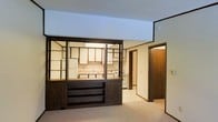 This single-floor space presents a notable open-concept design centered around a striking dark wood room divider that separates the living area from the kitchen. The divider features a combination of open shelving above and closed cabinetry drawers below, creating both visual interest and practical storage while maintaining sightlines throughout the space.

The kitchen is fully equipped with white appliances, including a refrigerator and range, complemented by cream-toned upper cabinetry arranged in an asymmetric pattern above a countertop with a built-in sink. The kitchen benefits from overhead fluorescent lighting, ensuring a well-lit workspace.

The living area is finished with neutral wall-to-wall carpeting, contributing to a warm, understated ambiance. A textured popcorn ceiling runs throughout, consistent with classic mid-century construction details. Dark wood trim frames the doorways and hallway, tying together the brown tones of the divider unit and adding visual cohesion.

A hallway visible from the main living area leads to additional rooms, suggesting a practical and straightforward floor plan. Overall, the space reflects a functional retro aesthetic with versatile layout potential.