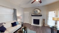 This single-floor space offers a thoughtfully designed living area that balances comfort and style. The focal point of the room is a classic white brick fireplace, complete with a dark mantel that provides a striking contrast and serves as an ideal display surface. An elegant oval mirror rests atop the mantel, enhancing the sense of depth and light in the room.

The flooring throughout is a rich, dark wood-style material that adds warmth and continuity to the space. A ceiling fan with dark wood-tone blades is centrally positioned, offering both air circulation and a subtle decorative element. The walls are finished in a soft neutral gray tone, providing a versatile backdrop that complements a range of décor styles.

Natural light filters in through a window dressed with patterned curtains in a blue and gray palette, adding texture and visual interest. The overall layout is open and efficiently arranged, allowing for a comfortable flow through the living area. The combination of the fireplace, warm flooring, and neutral palette creates an inviting and cozy ambiance that makes this a functional and welcoming living space.