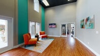 The University Village Club House in Carbondale, Illinois is a well-appointed community facility designed to serve residents with a variety of amenities under one roof. The main gathering area makes an immediate impression with soaring double-height ceilings, bold teal accent walls, and warm hardwood-style flooring that runs throughout the space. Large sliding glass doors and floor-to-ceiling windows flood the interior with natural light while offering direct views of the outdoor pool and patio area, creating a seamless connection between indoor and outdoor living.

The lounge area features vibrant orange accent chairs and a colorful area rug, providing a comfortable space to relax or socialize beneath a wall-mounted television. Adjacent to the lounge, a high-top dining table and bar seating offer a casual spot for gatherings, with the pool deck visible just beyond the expansive windows. A game room with a pool table adds a recreational element to the space.

The fitness center is well-equipped with cardio machines, strength equipment, wall-mounted televisions, and mirrors, all set beneath ample natural light from surrounding windows. A dedicated computer lounge provides workstations, high-top seating, and additional natural light — ideal for residents needing a productive workspace. A sleek reception desk with mosaic tile detailing serves as the welcoming point of contact for the facility.