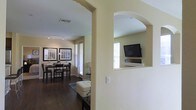 This single-story space showcases a thoughtfully designed open-concept layout, connecting the living and dining areas through a series of distinctive arched doorways that lend architectural character throughout. The warm, neutral-toned walls complement the rich, dark hardwood-style flooring, creating a cohesive and inviting ambiance.

The living area features a built-in media niche framed by an arched opening, providing a dedicated space for a flat-screen television and media components. This design element serves as a functional focal point while maintaining the smooth flow of the interior. Track lighting along the ceiling enhances illumination throughout the space.

The dining area is visible through the arched entryway, accommodating a full dining set beneath framed wall art that adds a polished, finished look. Multiple windows fitted with horizontal blinds allow natural light to filter in while offering privacy. The transition between spaces is seamless, with the open layout encouraging easy movement and interaction across the entire floor plan. The overall design balances practicality with a clean, well-appointed aesthetic.