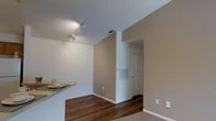 This single-floor space presents a clean, functional layout with a cohesive neutral color palette throughout. The main living area features rich, dark hardwood flooring that contrasts neatly with the warm beige-toned walls and white trim, creating a grounded, inviting atmosphere. Track lighting mounted on the ceiling provides directed illumination, adding a practical yet modern touch to the space.

A partial-height breakfast bar separates the kitchen area from the living space, offering a convenient casual dining or workspace surface. The open sightlines between these areas contribute to an efficient, connected feel. A centrally located HVAC vent on the wall ensures climate control is integrated unobtrusively into the design.

A hallway leads toward an interior door, suggesting additional rooms or storage beyond the main living area. The transition from hardwood to carpet is visible near the hallway, indicating a shift in flooring material in adjacent spaces. White walls and ceilings in the hallway area maintain a bright, clean aesthetic throughout the unit. Overall, the space balances practicality with a straightforward, well-maintained design.