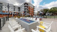 The Sun Deck at 28 Southfield Ave in Stamford, Connecticut is a well-appointed outdoor amenity space designed for relaxation and social gathering. At the heart of the deck is a rectangular, glass-enclosed gas fire pit set in a granite surround, serving as the centerpiece around which white Adirondack chairs are generously arranged for comfortable lounging. Accent seating in bold yellow adds a vibrant pop of color against the predominantly white palette of the space.

Toward the perimeter, a shaded bar area features blue-and-white striped market umbrellas and bright yellow bar stools, creating a lively and inviting spot for casual dining and entertaining. Additional colorful picnic-style tables are dispersed throughout the deck, offering versatile seating options.

Access to the Sun Deck is provided through a well-lit interior corridor finished with warm beige walls, patterned blue carpeting, and glass-panel double doors that frame views of the outdoor space. The corridor leads residents seamlessly from the building's interior to the open-air terrace.

The deck is open to the sky, offering sweeping views of the surrounding Stamford skyline and neighboring architecture. The combination of the fire pit, flexible seating arrangements, and thoughtfully designed access points makes this Sun Deck a functional and visually dynamic communal amenity.