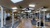 This fitness center is a single-floor facility designed to accommodate a full range of workout needs. The space features a vaulted ceiling that lends an open, airy feel, enhanced by multiple skylights that allow natural light to pour in throughout the day. Recessed and overhead fluorescent lighting ensures the space remains well-lit during all hours of operation.

The workout floor is finished with durable hardwood-style flooring complemented by protective rubber exercise mats positioned in key lifting zones. Full-length mirrors line the walls, making the space feel expansive while allowing users to monitor their form during exercise.

The equipment layout is thoughtfully organized, featuring a variety of strength-training machines, cable systems, power racks, and adjustable benches. Dedicated free weight areas provide ample space for a variety of exercises. Ceiling-mounted flat-screen televisions are strategically placed throughout the facility, providing entertainment and motivation during workouts.

A ceiling fan contributes to air circulation and comfort during intense training sessions. The overall ambiance is clean, functional, and professional, making it a well-rounded facility suited for users of all fitness levels.