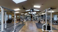 This fitness center is a single-floor facility designed to accommodate a full range of workout needs. The space features a vaulted ceiling that lends an open, airy feel, enhanced by multiple skylights that allow natural light to pour in throughout the day. Recessed and overhead fluorescent lighting ensures the space remains well-lit during all hours of operation.

The workout floor is finished with durable hardwood-style flooring complemented by protective rubber exercise mats positioned in key lifting zones. Full-length mirrors line the walls, making the space feel expansive while allowing users to monitor their form during exercise.

The equipment layout is thoughtfully organized, featuring a variety of strength-training machines, cable systems, power racks, and adjustable benches. Dedicated free weight areas provide ample space for a variety of exercises. Ceiling-mounted flat-screen televisions are strategically placed throughout the facility, providing entertainment and motivation during workouts.

A ceiling fan contributes to air circulation and comfort during intense training sessions. The overall ambiance is clean, functional, and professional, making it a well-rounded facility suited for users of all fitness levels.