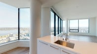 This high-rise residence showcases a contemporary design defined by clean lines, a neutral white palette, and light-toned hardwood flooring throughout. The open-concept living and kitchen area is the centerpiece of the space, wrapped in expansive floor-to-ceiling windows on multiple sides that frame breathtaking panoramic views of the city skyline, a waterway, and surrounding neighborhoods. Natural light floods every corner, reinforcing the unit's bright, airy character.

The kitchen is outfitted with sleek white cabinetry featuring brushed metal hardware, a stainless steel undermount sink, gas cooktop, and full-size stainless steel refrigerator. A generous white quartz peninsula provides additional workspace and a natural transition into the open living area.

Multiple bedrooms offer hardwood flooring and large windows with commanding elevated views. One bedroom connects directly to a dedicated bathroom, while others share access to additional facilities. The primary bathroom is particularly well-appointed, featuring a dual-sink white quartz vanity, a soaking tub, and a glass-enclosed tiled shower. A secondary bathroom includes a walk-in glass shower with tile surround and mosaic flooring, while a third bathroom offers a clean, minimalist vanity layout with tile flooring.