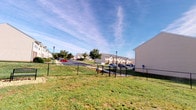 This single-floor residential space is situated within a well-established neighborhood community characterized by neatly maintained green lawns and a welcoming outdoor environment. The exterior grounds feature a chain-link fenced area that defines the property boundary while preserving open sightlines to the surrounding community. A classic wooden bench is positioned within the fenced grounds, offering a dedicated spot for outdoor relaxation and leisure. Mature trees line the perimeter and neighboring areas, providing natural shade and a lush backdrop throughout the warmer months. Street lamps are stationed along the sidewalks and pathways, ensuring well-lit circulation through the community at all hours. The surrounding neighborhood consists of multi-story townhomes with neutral-toned vinyl siding, contributing to a cohesive residential aesthetic. Paved parking areas are conveniently accessible, accommodating multiple vehicles for residents and guests. The open sky and expansive lawn areas give the community a spacious, airy feel, while the combination of greenery, structured fencing, and community amenities creates a balanced environment suited for everyday living. Overall, the outdoor setting reflects a functional and pleasant residential community designed with both practicality and comfort in mind.