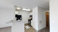 This single-floor space showcases a thoughtfully designed, modern interior with a clean and functional aesthetic throughout. The kitchen is a central highlight, featuring crisp white cabinetry that extends to the ceiling, providing ample storage and a bright, airy feel. Stainless steel appliances — including a gas range, built-in microwave, and a full-size refrigerator — lend a sleek, contemporary look to the cooking area. A peninsula counter extends into the adjacent space, creating a natural division between the kitchen and the living area while offering additional prep and casual dining space. The countertops feature a light stone-like finish that complements the white cabinetry and neutral wall tones. The flooring consists of durable wood-look planks in a warm tone, running throughout the space and adding a grounding contrast to the bright white surroundings. Recessed and flush-mounted ceiling lighting ensures the kitchen is well-illuminated at all times. The overall ambiance is clean, minimal, and functional, with a neutral palette that offers a versatile canvas for personalization. The layout is efficient and well-organized, making excellent use of the available space.