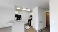 This single-floor space showcases a thoughtfully designed, modern interior with a clean and functional aesthetic throughout. The kitchen is a central highlight, featuring crisp white cabinetry that extends to the ceiling, providing ample storage and a bright, airy feel. Stainless steel appliances — including a gas range, built-in microwave, and a full-size refrigerator — lend a sleek, contemporary look to the cooking area. A peninsula counter extends into the adjacent space, creating a natural division between the kitchen and the living area while offering additional prep and casual dining space. The countertops feature a light stone-like finish that complements the white cabinetry and neutral wall tones. The flooring consists of durable wood-look planks in a warm tone, running throughout the space and adding a grounding contrast to the bright white surroundings. Recessed and flush-mounted ceiling lighting ensures the kitchen is well-illuminated at all times. The overall ambiance is clean, minimal, and functional, with a neutral palette that offers a versatile canvas for personalization. The layout is efficient and well-organized, making excellent use of the available space.