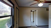 This two-story space presents a straightforward and functional layout. The interior is characterized by neutral, light-colored walls contrasted by dark baseboards and door trim, creating a clean, defined aesthetic throughout. Carpeted flooring runs through the rooms, adding warmth and comfort underfoot.

The room visible in the image features a ceiling fan with a brass-toned fixture, providing both air circulation and overhead lighting. A window along one wall allows natural light to filter into the space, brightening the interior. Dark-trimmed doorways connect the rooms, with an adjoining space visible beyond — appearing to include a closet or storage area, offering practical organization solutions.

The layout suggests an open flow between rooms, with doorways that create a connected feel across the floor plan. The ceiling height appears standard, contributing to a comfortable and proportionate sense of space. The overall ambiance is modest and neutral, providing a blank canvas that can accommodate a variety of furnishing styles and functional uses. The two-floor configuration further adds versatility to the overall space.
