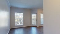 This single-story space is designed with a clean, functional layout that makes efficient use of its footprint. The living area serves as the heart of the unit, finished with dark wood-look flooring and elegant crown molding along the ceiling perimeter. Multiple large windows and a glass-paneled exterior door flood the room with natural light while framing views of surrounding mature trees, creating a bright and airy atmosphere. The exterior door opens to what appears to be a balcony or patio, adding an outdoor element to the living space.

The kitchen is compact and practical, featuring laminate countertops with wood-trimmed edges, white cabinetry with wood accents, and an electric coil range with an overhead exhaust hood, providing everything needed for everyday cooking.

The bathroom is well-appointed with a single-sink vanity topped with matching laminate counters, white cabinetry beneath, a large wall mirror with overhead lighting, and a tiled shower-tub combination. Wood-look flooring carries through this space as well.

The bedroom is a comfortable, well-lit room enhanced by a trio of windows offering treetop views, a ceiling fan with light fixture, and the same wood-look flooring found throughout the unit. Neutral wall tones throughout contribute to a cohesive, calm overall ambiance.