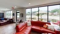 This single-floor space is thoughtfully designed to maximize natural light and openness. The living area is anchored by large floor-to-ceiling windows and a sliding glass door that lead to an outdoor balcony equipped with patio furniture and umbrellas, offering expansive outdoor views. Two distinct seating areas are present: one featuring a bold red leather sectional sofa accented with decorative pillows, and another with a generously sized brown sectional, each complemented by glass accent tables and nature-inspired wall art. Recessed lighting and stone tile flooring run throughout, contributing to a clean, contemporary aesthetic.

The kitchen is well-appointed with rich cherry wood cabinetry, dark granite countertops, and a classic tile backsplash. Stainless steel appliances, including a full-size refrigerator, microwave, and dishwasher, provide full functionality. A peninsula with dark granite countertops extends into the space, offering additional seating with vibrant teal and neutral bar stools. A large window above the peninsula frames views of mature trees, further connecting the interior to its natural surroundings. The overall ambiance of the space is modern yet inviting, blending warm tones with contemporary design elements.