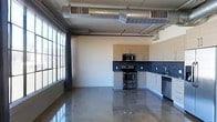 This single-floor space showcases a striking industrial-modern aesthetic that blends raw architectural elements with contemporary finishes. The open-plan layout is anchored by gleaming polished concrete floors that reflect light beautifully throughout the space, lending a sleek, expansive feel underfoot.

The kitchen is a standout feature, outfitted with stainless steel appliances including a range with oven, built-in microwave, and dishwasher. Light wood-toned cabinetry lines the walls in an L-shaped configuration, complemented by a bold deep-blue mosaic tile backsplash that adds texture and visual interest. The clean lines of the cabinetry and modern hardware contribute to the overall contemporary character of the space.

Overhead, exposed silver ductwork and conduit run prominently across the ceiling, reinforcing the industrial design language and adding architectural drama. The elevated ceiling height enhances the sense of openness and volume throughout the unit.

Large industrial-style windows along one wall flood the space with generous natural light, while floor-length curtains provide the option for privacy. The combination of raw materials, modern fixtures, and abundant light creates an ambiance that is both functional and distinctly urban in character.
