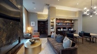 Located at 225 S Grand Ave in the heart of downtown Los Angeles, this single-floor amenity lounge is a thoughtfully designed communal space that blends functionality with upscale interior design. The space is organized into several distinct zones, each serving a specific purpose while maintaining a cohesive, contemporary aesthetic throughout.

A dedicated business center features a built-in walnut desk spanning the width of a recessed alcove, accommodating multiple workstations with desktop computers. Open shelving above displays an organized collection of books and decorative objects, lending a scholarly atmosphere. Nearby, round light-wood tables with leather-upholstered chairs provide additional casual work or meeting areas.

A lounge seating area is anchored by a dramatic floor-to-ceiling fireplace clad in richly veined dark stone, with a wall-mounted television above. Floor-to-ceiling sheer drapery frames expansive windows that offer views of mature trees outdoors, filling the space with natural light. Plush sofas, leather armchairs, and layered area rugs create a warm, inviting atmosphere.

A library nook features a full wall of built-in bookshelves curating colorful volumes alongside decorative accents, surrounded by club chairs arranged around a low coffee table. Overhead, a sculptural multi-globe chandelier adds an artistic focal point. Recessed lighting and elevated ceilings throughout contribute to the space's open, refined ambiance.