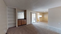 This single-floor space offers an open and functional layout designed to maximize usability. The main living area is finished with wall-to-wall carpeting in a neutral tone, creating a warm and understated foundation throughout the room. A textured popcorn ceiling adds dimension overhead, while recessed lighting provides focused illumination in select areas.

A standout feature of the living space is the built-in wet bar, complete with wood cabinetry, a dark countertop, and a mirrored backsplash that reflects light and adds a sense of depth to the room. Adjacent to the wet bar is a set of open, floor-to-ceiling shelving units, offering versatile display or storage options.

Natural light enters the space through a sliding glass door, which provides direct access to an outdoor patio area, visually connecting the interior to the exterior. On the opposite side of the living area, an opening leads into the kitchen, which features light-colored cabinetry and appears to connect seamlessly to the rest of the layout.

Overall, the space presents a straightforward, open design with practical built-in features and easy indoor-outdoor flow.