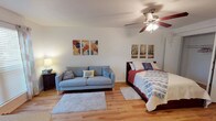This single-floor, one-bedroom, one-bathroom unit offers a thoughtfully designed open-concept layout that maximizes every square foot of its compact footprint. The main living area seamlessly integrates the sleeping and lounging zones, creating a flexible and functional space. Warm hardwood-style flooring runs throughout, lending a clean, cohesive aesthetic to the interior.

A ceiling fan with an integrated light fixture is centrally positioned overhead, providing both air circulation and ambient lighting. Natural light filters into the space through a window dressed with blinds and decorative curtains, brightening the room and contributing to an airy atmosphere.

The sleeping area is well-appointed and benefits from generous storage, featuring two side-by-side reach-in closets with built-in shelving and hanging rods — a practical and notable feature for a space of this size. A partial view of the kitchenette reveals white cabinetry and a breakfast bar-style counter, offering a dedicated space for meal preparation and casual dining.

The neutral color palette of white walls paired with the warm flooring tones creates a bright and welcoming ambiance throughout. Overall, this unit is a well-organized, efficiently laid-out space suited for comfortable single-occupant living.