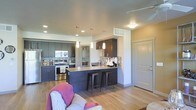 This single-floor residence showcases a thoughtfully designed open-concept layout that integrates the kitchen and living areas into one cohesive, functional space. The kitchen is a standout feature, outfitted with dark espresso-toned cabinetry that extends to the upper walls, providing generous storage capacity. A mosaic tile backsplash adds texture and visual interest, complementing the clean lines of the stainless steel appliances, including a range and built-in microwave. A glass-front display cabinet offers an elegant accent to the cabinetry arrangement. The kitchen peninsula serves as a natural dividing element between the cooking and living zones, featuring a dark countertop and seating for multiple guests via industrial-style bar stools. Pendant lighting in varying heights hangs above the peninsula, adding warmth and a contemporary aesthetic. Recessed lighting throughout ensures the space is well-lit. Warm, light-toned hardwood-style flooring runs continuously across the entire floor, unifying the open layout. The entry door is positioned conveniently off the main living area, and the overall ambiance is modern, clean, and welcoming.
