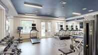Located at 110 Talisman Way in Raleigh, NC, this fitness center is a comprehensively outfitted exercise facility spread across a single floor. The space is divided into distinct workout zones, each designed to support a full range of training styles.

The main cardio and strength area features a bold blue ceiling with recessed and linear lighting that creates a bright, energizing atmosphere. Large windows and glass French doors flood the space with natural light and offer views of an outdoor pool area. Treadmills and elliptical machines equipped with individual monitors line one wall, complemented by a full cable machine system and weight benches for strength training. A wall-length mirror expands the sense of space and allows for form monitoring. A dumbbell rack rounds out the free-weight offerings.

A dedicated functional training room features rich dark hardwood flooring, a dark ceiling with recessed lighting, and a more intimate feel. This area is stocked with kettlebells, medicine balls, stability balls, TRX suspension trainers, resistance bands, and a flat-screen TV, making it ideal for dynamic and group-style workouts.

The flooring throughout the primary zone is durable speckled rubber, well-suited for high-traffic fitness use. A WaterRower rowing machine adds a premium cardio option. The overall design blends a clean, modern aesthetic with a functional layout, providing a professional-grade fitness experience.