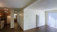 This single-floor residence offers a straightforward and functional layout designed for comfortable everyday living. The main living area features dark hardwood-style flooring that extends throughout the space, providing a warm and cohesive look underfoot. The textured popcorn ceiling adds a classic residential character to the rooms. A wall-mounted heater unit is conveniently positioned in the main living area, serving as the primary heat source for the space.

A hallway connects the living area to the rest of the home, leading toward what appears to be a bathroom, which is visible in the background. The bathroom is equipped with a vanity featuring warm wood-toned cabinetry and a well-lit mirror, illuminated by a row of vanity lights above. The neutral white walls throughout create a clean, bright atmosphere that allows for versatile interior styling.

The overall layout is efficiently organized, with each area flowing naturally into the next. The combination of dark flooring, white walls, and warm wood accents in the bathroom gives the space a balanced, neutral aesthetic that would suit a variety of furnishing styles. The single-floor design ensures easy accessibility and convenient navigation throughout the home.