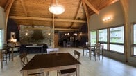 This single-floor communal space combines rustic charm with functional amenities, making it an inviting venue for gatherings and events. The most striking architectural feature is the soaring vaulted ceiling clad in warm natural wood planking, supported by exposed timber beams and arched trusses that create a grand, lodge-like atmosphere throughout the open interior.

A prominent stone-clad fireplace with a large custom metal hood anchors one side of the space, adding both a visual focal point and a cozy, rustic character. The open-concept layout flows seamlessly from the dining area into a well-equipped kitchen, which features dark cabinetry, modern appliances including a full-size refrigerator, and pendant lighting over a raised bar counter with seating for several guests.

Multiple dining tables with wooden chairs are arranged throughout the space, accommodating a variety of group sizes. Light-toned tile flooring runs throughout, complementing the warm wood tones above. Large windows and glass doors along one wall flood the interior with natural light and offer views of the surrounding greenery, creating a bright and welcoming ambiance that balances the darker, earthy design elements. A decorative globe pendant light fixture hangs centrally, adding an elegant finishing touch to this well-appointed space.