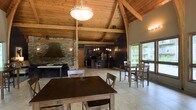 This single-floor communal space combines rustic charm with functional amenities, making it an inviting venue for gatherings and events. The most striking architectural feature is the soaring vaulted ceiling clad in warm natural wood planking, supported by exposed timber beams and arched trusses that create a grand, lodge-like atmosphere throughout the open interior.

A prominent stone-clad fireplace with a large custom metal hood anchors one side of the space, adding both a visual focal point and a cozy, rustic character. The open-concept layout flows seamlessly from the dining area into a well-equipped kitchen, which features dark cabinetry, modern appliances including a full-size refrigerator, and pendant lighting over a raised bar counter with seating for several guests.

Multiple dining tables with wooden chairs are arranged throughout the space, accommodating a variety of group sizes. Light-toned tile flooring runs throughout, complementing the warm wood tones above. Large windows and glass doors along one wall flood the interior with natural light and offer views of the surrounding greenery, creating a bright and welcoming ambiance that balances the darker, earthy design elements. A decorative globe pendant light fixture hangs centrally, adding an elegant finishing touch to this well-appointed space.