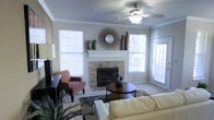 This single-floor living space centers around a tile-surround fireplace with a crisp white mantel, serving as the focal point of the main living area. The fireplace is framed by decorative vases and a circular mirror, adding visual interest to the space. Crown molding lines the perimeter of the room, contributing a refined architectural detail that elevates the overall aesthetic.

The living area is well-lit, benefiting from multiple windows fitted with horizontal blinds, allowing for adjustable natural light throughout the day. A glass-paneled door further enhances the brightness and provides access to an adjacent area or exterior. Neutral gray walls create a versatile backdrop that complements both the light-toned carpet and the varied furnishings.

A ceiling fan with an integrated light fixture provides both comfort and ambient lighting. The room comfortably accommodates a sectional sofa, accent chair, and a round coffee table atop a patterned area rug, demonstrating a practical and inviting layout. Overall, the space offers a warm, functional atmosphere suited for everyday living and entertaining.
