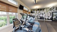 This fitness center offers a comprehensive and thoughtfully designed workout environment suited for a wide range of exercise routines. The space is anchored by a striking full-wall mural depicting a forest scene with abstract white orbs, lending an artistic and energizing ambiance to the room. Durable rubber flooring runs throughout, providing safety and comfort underfoot across all workout zones.

The cardio area is generously outfitted with a row of Precor treadmills, elliptical cross-trainers, and a stationary bike, each positioned to take advantage of the abundant natural light streaming through large windows fitted with adjustable roller shades. Wall-mounted flat-screen televisions are placed throughout for entertainment during workouts.

The strength training section features multiple cable and resistance machines alongside a well-stocked dumbbell rack, catering to a full range of muscle groups. Large mirrors line one wall, enhancing the sense of openness while allowing users to monitor their form.

Ceiling-mounted and wall-mounted fans provide ventilation, while recessed lighting ensures the space remains bright. A sanitizing station near the entrance promotes cleanliness. The overall layout is organized and spacious, creating a clean, modern fitness environment that balances functionality with an inviting aesthetic.