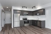 This thoughtfully designed single-floor space combines contemporary finishes with a clean, minimalist aesthetic. Warm wood-look flooring flows consistently throughout, tying each area together seamlessly. A hallway with a sleek sliding barn door provides a stylish entry point, leading into the main living areas.

The open living space is a standout feature, bathed in natural light from multiple large windows that frame views of the surrounding neighborhood. The kitchen is well-equipped with stainless steel appliances including a refrigerator, range, microwave, and dishwasher. Two-tone cabinetry — pairing rich wood uppers with darker lower cabinets — is complemented by crisp white countertops and track lighting overhead, creating a modern, functional workspace.

The bedrooms are bright and airy, each featuring multiple windows with roller shades that let in generous daylight and offer outdoor views. One room includes a wall-mounted mini-split climate control unit for personalized comfort, while another opens to a private outdoor area through a full-length sliding glass door. A well-organized closet with built-in shelving and hanging rods provides ample storage.

The bathroom features a clean, white tub and shower combination with chrome fixtures. Throughout the space, white walls and recessed and flush lighting maintain a bright, welcoming ambiance.