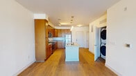 This single-floor residence offers a thoughtfully designed open-concept layout that seamlessly connects the kitchen and living areas. The kitchen is a standout feature, showcasing warm honey-toned cabinetry that reaches toward the ceiling, providing generous storage throughout. Granite countertops extend across the perimeter and onto a central island, which includes an undermount sink and serves as both a workspace and casual gathering spot. Stainless steel appliances — including a refrigerator, range, and built-in microwave — complete the kitchen's functional and polished aesthetic. Pendant lighting above the island adds a refined touch. Hardwood flooring flows consistently throughout the main living areas, lending warmth and continuity to the space. The living room is bright and open, with large sliding glass doors dressed with vertical blinds that invite natural light and offer views to the outdoors. A dedicated in-unit laundry closet with a stacked washer and dryer adds everyday convenience. Two bathrooms are finished with granite-topped vanities and warm wood cabinetry; one features a full tub-shower combination with classic white tile surround, while the other is appointed as a half bath. Light-toned tile flooring in the bathrooms complements the overall palette of the residence.