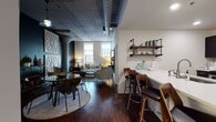 Located at 1215 W Tenth St in Cleveland's vibrant Ohio City neighborhood, this single-floor loft residence masterfully combines industrial heritage with contemporary design. The open-plan living and dining area anchors the space, featuring rich hardwood floors, exposed concrete columns, and overhead ductwork that pay homage to the building's industrial roots. Decorative lattice wall panels add an artistic focal point, while the living room benefits from natural light streaming through large windows with views of the surrounding cityscape.

The kitchen is outfitted with dark cabinetry, stainless steel appliances, light countertops, and industrial-style pipe shelving — a practical yet visually striking workspace. A breakfast bar with stylish seating connects seamlessly to the dining area, enhancing the open flow of the space.

The residence includes multiple bedrooms, each with its own distinct character. One bedroom showcases a dramatic reclaimed wood accent wall with warm underlighting, while another offers a serene, light-filled retreat with tall ceilings and soft carpeting. The bathroom features a clean, modern aesthetic with dark vanity cabinetry, a lighted mirror, and tiled flooring. Throughout, the space strikes a refined balance between urban industrial character and comfortable, well-appointed living.