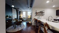 Located at 1215 W Tenth St in Cleveland's vibrant Ohio City neighborhood, this single-floor loft residence masterfully combines industrial heritage with contemporary design. The open-plan living and dining area anchors the space, featuring rich hardwood floors, exposed concrete columns, and overhead ductwork that pay homage to the building's industrial roots. Decorative lattice wall panels add an artistic focal point, while the living room benefits from natural light streaming through large windows with views of the surrounding cityscape.

The kitchen is outfitted with dark cabinetry, stainless steel appliances, light countertops, and industrial-style pipe shelving — a practical yet visually striking workspace. A breakfast bar with stylish seating connects seamlessly to the dining area, enhancing the open flow of the space.

The residence includes multiple bedrooms, each with its own distinct character. One bedroom showcases a dramatic reclaimed wood accent wall with warm underlighting, while another offers a serene, light-filled retreat with tall ceilings and soft carpeting. The bathroom features a clean, modern aesthetic with dark vanity cabinetry, a lighted mirror, and tiled flooring. Throughout, the space strikes a refined balance between urban industrial character and comfortable, well-appointed living.