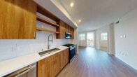 This well-appointed single-floor residence showcases a thoughtfully designed open layout that seamlessly connects the kitchen and living areas. The kitchen is a standout feature, outfitted with warm wood cabinetry, open shelving, and a white subway tile backsplash that provides a clean, modern contrast. A full suite of stainless steel appliances — including a gas range, built-in microwave, dishwasher, and French door refrigerator — completes the space. White quartz countertops and a deep single-basin sink with a contemporary pull-down faucet add both function and style. Light hardwood-style flooring flows throughout the main living areas, enhancing the bright, airy atmosphere created by recessed lighting and large windows that invite natural light. The bedroom is a calm, neutral space featuring plush carpeting and a window with outdoor views. The bathroom is elegantly finished with large-format marble-look tile on both the floors and shower surround, a glass-enclosed tub-shower combo, and clean white walls. A walk-in closet is conveniently accessible directly from the bathroom. The overall design balances contemporary finishes with a warm, livable aesthetic throughout.