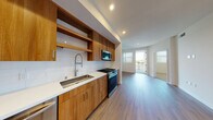 This well-appointed single-floor residence showcases a thoughtfully designed open layout that seamlessly connects the kitchen and living areas. The kitchen is a standout feature, outfitted with warm wood cabinetry, open shelving, and a white subway tile backsplash that provides a clean, modern contrast. A full suite of stainless steel appliances — including a gas range, built-in microwave, dishwasher, and French door refrigerator — completes the space. White quartz countertops and a deep single-basin sink with a contemporary pull-down faucet add both function and style. Light hardwood-style flooring flows throughout the main living areas, enhancing the bright, airy atmosphere created by recessed lighting and large windows that invite natural light. The bedroom is a calm, neutral space featuring plush carpeting and a window with outdoor views. The bathroom is elegantly finished with large-format marble-look tile on both the floors and shower surround, a glass-enclosed tub-shower combo, and clean white walls. A walk-in closet is conveniently accessible directly from the bathroom. The overall design balances contemporary finishes with a warm, livable aesthetic throughout.