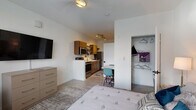 Located at 180 N Martin Luther King Blvd in downtown Lexington, Kentucky, this studio unit offers a thoughtfully designed open-concept layout that seamlessly integrates the living, sleeping, and kitchen areas into one cohesive space. Light-toned luxury vinyl plank flooring runs throughout, lending a clean and modern aesthetic.

The sleeping area is well-appointed with a ceiling fan, a large window dressed with horizontal blinds that invites natural light, and a wall-mounted flat-screen television paired with a multi-drawer dresser for convenient storage. A dedicated walk-in closet with built-in shelving and hanging rods provides generous organization space.

The kitchen is efficiently equipped with warm wood cabinetry, stainless steel appliances including a full-size refrigerator, microwave, and range, as well as a deep sink with a modern faucet and open metal shelving for additional storage. A small dining area with seating sits just adjacent, making everyday meals practical and comfortable.

The full bathroom features warm wood cabinetry beneath a white countertop vanity, a large mirror with bright overhead lighting, a tub and shower combination with a curtain, and tasteful flooring details. The overall ambiance of the unit is bright, clean, and functional, making it well-suited for single-occupant urban living in the heart of Lexington.