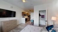 Located at 180 N Martin Luther King Blvd in downtown Lexington, Kentucky, this studio unit offers a thoughtfully designed open-concept layout that seamlessly integrates the living, sleeping, and kitchen areas into one cohesive space. Light-toned luxury vinyl plank flooring runs throughout, lending a clean and modern aesthetic.

The sleeping area is well-appointed with a ceiling fan, a large window dressed with horizontal blinds that invites natural light, and a wall-mounted flat-screen television paired with a multi-drawer dresser for convenient storage. A dedicated walk-in closet with built-in shelving and hanging rods provides generous organization space.

The kitchen is efficiently equipped with warm wood cabinetry, stainless steel appliances including a full-size refrigerator, microwave, and range, as well as a deep sink with a modern faucet and open metal shelving for additional storage. A small dining area with seating sits just adjacent, making everyday meals practical and comfortable.

The full bathroom features warm wood cabinetry beneath a white countertop vanity, a large mirror with bright overhead lighting, a tub and shower combination with a curtain, and tasteful flooring details. The overall ambiance of the unit is bright, clean, and functional, making it well-suited for single-occupant urban living in the heart of Lexington.