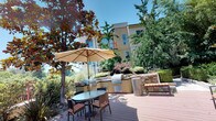This inviting outdoor deck space offers a well-appointed communal retreat set within a lush, tree-lined environment. The expansive wooden deck serves as the central gathering area, thoughtfully designed with multiple seating zones to accommodate both relaxation and dining. A prominent stone-framed raised planter anchors the center of the deck, adding a decorative architectural element and a touch of natural charm to the space.

Scattered across the deck are comfortable lounge chairs with cushioned seating, as well as a shaded dining set positioned beneath a large market umbrella, ideal for outdoor meals in a comfortable setting. The space is enveloped by mature trees and well-maintained landscaping, creating a sense of privacy and tranquility despite its communal nature.

One of the standout features of this outdoor space is the fully equipped outdoor kitchen area, complete with stainless steel grills, counter workspace, and built-in cabinetry finished with stone detailing. An additional shaded dining table is positioned nearby, making the kitchen zone perfect for al fresco entertaining. The warm, sun-drenched atmosphere, open sky views, and surrounding greenery contribute to an overall ambiance that is both serene and functional — a true outdoor living destination.