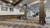 This fitness center is a thoughtfully designed workout facility that blends upscale aesthetics with comprehensive exercise amenities. The space is anchored by a distinctive hexagonal-patterned carpet in neutral tones of gray and taupe, complemented by decorative columns with paneled detailing that define separate workout zones while maintaining an open, airy feel.

The cardio zone is generously equipped with treadmills, ellipticals, and stationary bikes, all positioned near large windows and glass doors that invite abundant natural light and offer views of the outdoor surroundings. Full-length mirrors along the walls amplify the sense of space and allow exercisers to monitor their form. Ceiling fans provide additional airflow throughout.

A dedicated free weights area features a full rack of dumbbells alongside wall-mounted mirrors, framed windows, and wainscoting details that add a refined architectural touch. The functional training zone stands out with a multi-use rig equipped with TRX suspension trainers, a heavy punching bag, pull-up bars, and barbell storage — catering to a wide range of workout styles.

The entry foyer features an elegant tiled floor and tall glass-paneled doors that open to an outdoor area, creating a seamless indoor-outdoor connection. Recessed lighting throughout ensures consistent, well-lit conditions across all workout areas, making this fitness center a polished and fully equipped space for daily training.