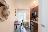 This single-floor studio is thoughtfully designed to maximize functionality within a compact footprint. The entry welcomes residents through a classic six-panel white door, complemented by neutral warm-toned walls and dark tile flooring that set a clean, understated tone throughout.

The main living area seamlessly integrates sleeping, studying, and cooking into one cohesive space. A built-in kitchenette lines one wall, featuring rich dark-wood cabinetry, granite-style countertops, a microwave, and a compact refrigerator — providing all the essentials for everyday living. Adjacent to the kitchenette, a wooden desk and chair offer a dedicated workspace, ideal for studying or remote work.

The sleeping area is positioned near a set of full-length French-style doors framed by horizontal blinds, allowing abundant natural light to filter in while offering views of lush greenery outside. The bedroom nook is cozy and well-appointed, with ample space for a twin bed and nearby storage solutions including shelving and woven baskets. Concrete-style flooring runs throughout, adding an industrial-modern touch that contrasts warmly with the soft, neutral wall tones. Overall, the space feels bright, practical, and efficiently organized for comfortable single-occupant living.