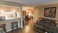 This single-floor space is designed with an open, flowing layout that seamlessly connects the kitchen, dining, and living areas. The rich hardwood flooring runs throughout, adding warmth and a cohesive feel to the entire space. Warm beige and tan wall tones complement the dark wood accents found in the furniture and architectural details.

The kitchen features a pass-through counter with bar-height seating, providing a practical and social connection to the adjacent living area. The counter appears to offer granite or stone-style surfaces, adding a refined touch.

The dining area is anchored by an elegant chandelier that casts a warm glow over the dining table and cushioned chairs. Framed artwork on the walls adds character to the space.

The living area is comfortably appointed with a plush sofa, decorative accent pillows, and a glass-top coffee table. A framed piece of wall art provides a bold focal point. A doorway visible from the living area suggests access to additional private rooms beyond. Overall, the space offers a welcoming and well-coordinated atmosphere suitable for both everyday living and entertaining.