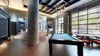 Located at 611 S Charles Street in Baltimore, MD, this contemporary clubhouse delivers an elevated amenity experience through thoughtful design and high-end finishes. The space is anchored by soaring coffered ceilings dressed in dark tones, dramatic pendant lighting, and substantial cylindrical concrete columns that lend an industrial-chic character throughout.

The main lounge area serves as the social heart of the clubhouse, featuring a sweeping curved sectional sofa, designer accent chairs, and a stunning cascading gold-and-crystal chandelier that draws the eye upward. A textured geometric feature wall frames a wall-mounted flat-screen television above a sleek linear fireplace, creating a compelling focal point. Large steel-framed windows flood the space with natural light and offer views of the surrounding greenery.

A dedicated billiards area with rich hardwood flooring and an industrial-style pendant chandelier provides a refined space for recreation. An entertainment zone includes a wet bar with dark cabinetry, mosaic tile backsplash, and an additional mounted television, ideal for gatherings.

The entry corridor features dramatic stone tile flooring, sculpted wall cladding, and a striking mirrored art installation with built-in bench seating. Adjacent, a glass-enclosed fitness center is fully equipped with modern exercise machines, maintaining visual connectivity to the rest of the clubhouse.