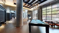 Located at 611 S Charles Street in Baltimore, MD, this contemporary clubhouse delivers an elevated amenity experience through thoughtful design and high-end finishes. The space is anchored by soaring coffered ceilings dressed in dark tones, dramatic pendant lighting, and substantial cylindrical concrete columns that lend an industrial-chic character throughout.

The main lounge area serves as the social heart of the clubhouse, featuring a sweeping curved sectional sofa, designer accent chairs, and a stunning cascading gold-and-crystal chandelier that draws the eye upward. A textured geometric feature wall frames a wall-mounted flat-screen television above a sleek linear fireplace, creating a compelling focal point. Large steel-framed windows flood the space with natural light and offer views of the surrounding greenery.

A dedicated billiards area with rich hardwood flooring and an industrial-style pendant chandelier provides a refined space for recreation. An entertainment zone includes a wet bar with dark cabinetry, mosaic tile backsplash, and an additional mounted television, ideal for gatherings.

The entry corridor features dramatic stone tile flooring, sculpted wall cladding, and a striking mirrored art installation with built-in bench seating. Adjacent, a glass-enclosed fitness center is fully equipped with modern exercise machines, maintaining visual connectivity to the rest of the clubhouse.