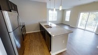 This single-floor residence showcases a thoughtfully designed open-concept layout that seamlessly connects the kitchen and living areas. The kitchen serves as the centerpiece of the space, featuring a large island with striking granite countertops and an undermount sink, complemented by dark cabinetry that provides a bold contrast to the light stone surfaces. Stainless steel appliances add a sleek, modern touch, while pendant lighting suspended above the island creates an inviting focal point. Warm hardwood-style flooring flows consistently throughout, unifying the kitchen and the adjacent living and dining areas. The open living space is bright and airy, benefiting from multiple windows that allow generous natural light to fill the room, creating a welcoming and comfortable atmosphere. Neutral gray-toned walls provide a versatile backdrop, lending the space a clean, contemporary aesthetic that adapts easily to a variety of interior styles. The overall design prioritizes functionality and flow, making this an ideal space for both everyday living and entertaining.